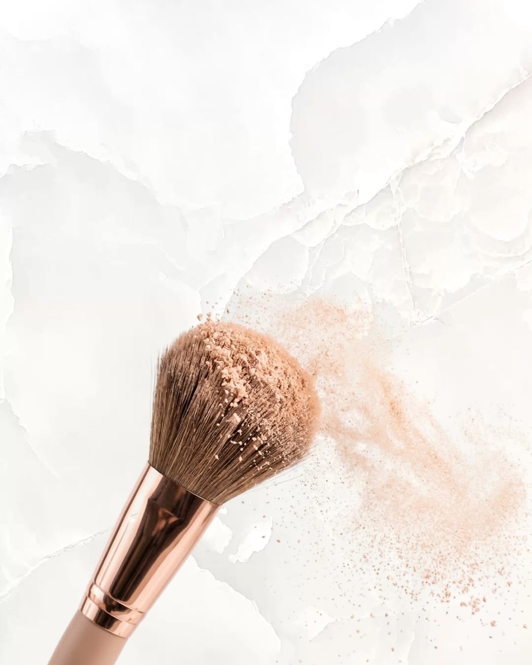 A rose-gold makeup brush with a cloud of powder on white marble